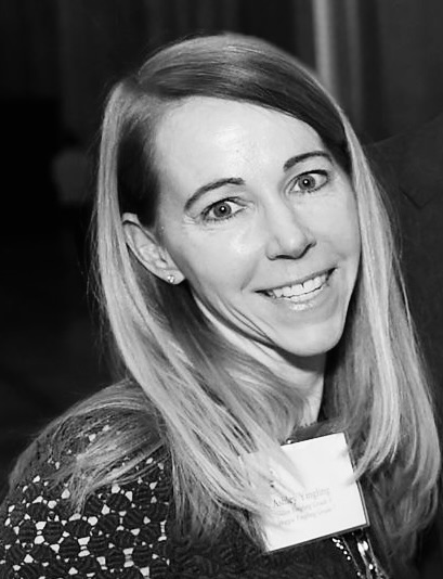 A woman with long, straight hair smiles at the camera while wearing a patterned top and a name tag. The photo is black and white.